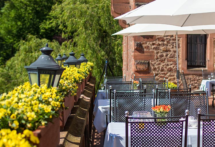 Die Außenterrasse des Restaurants im Schloss Saareck ist mit Metallstühlen und Tischen ausgestattet, die mit weißen Tischdecken bedeckt sind und deren Ränder mit leuchtend gelben Blumen geschmückt sind. Sonnenschirme spenden Schatten vor der sanften Brise der Saar, während im Hintergrund eine charmante Backsteinmauer mit Fenstern steht.