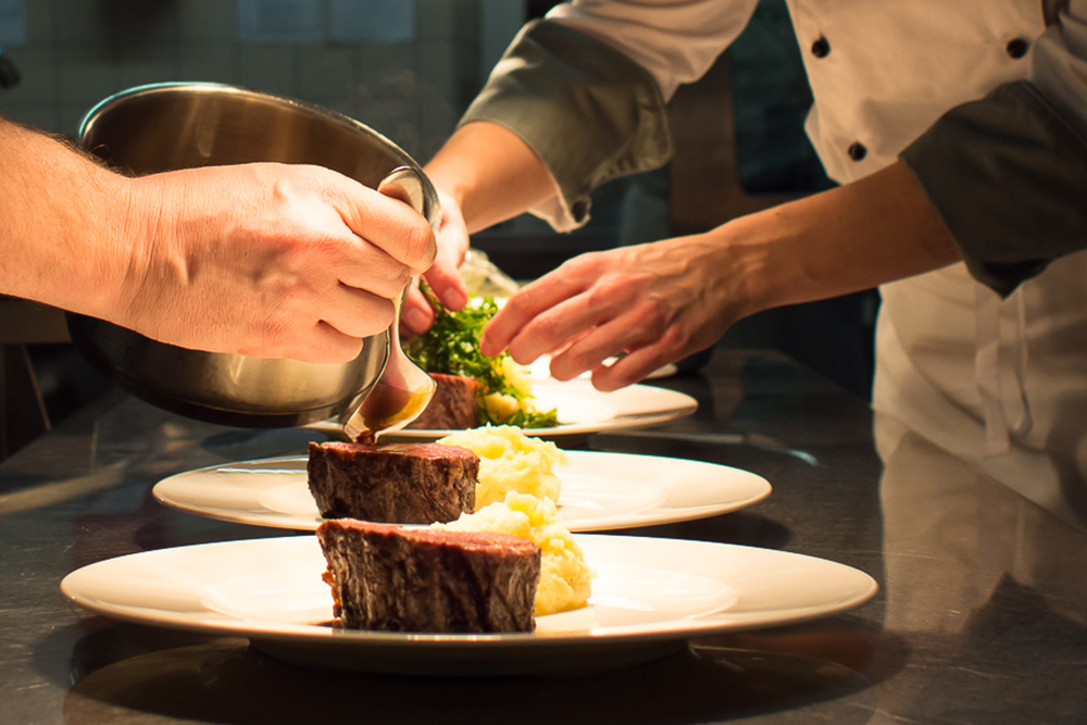 Zwei Köche richten ein Gericht mit einer Scheibe Fleisch, Kartoffelbrei und Gemüse an. Ein Koch gießt Soße über das Fleisch, während der andere das Essen auf einem weißen Teller anrichtet.