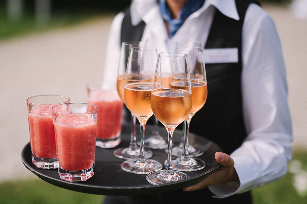 Der Kellner hält ein Tablett mit Gläsern rosafarbener Getränke; einige sind mit einem Mixgetränk gefüllt, andere mit einem Sprudelgetränk.