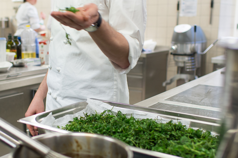 Koch hält in einer Großküche Kräuter über einem Tablett mit Gemüse.