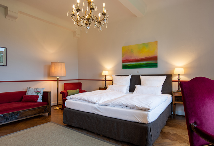 Ein Schlafzimmer, das Gemütlichkeit ausstrahlt, verfügt über ein Doppelbett, zwei Nachttischlampen, einen roten Sessel, ein rotes Sofa und eine große Lampe. Ein farbenfrohes Gemälde ergänzt den Kronleuchter an der Decke und verkörpert zeitlose Eleganz der Baukultur.