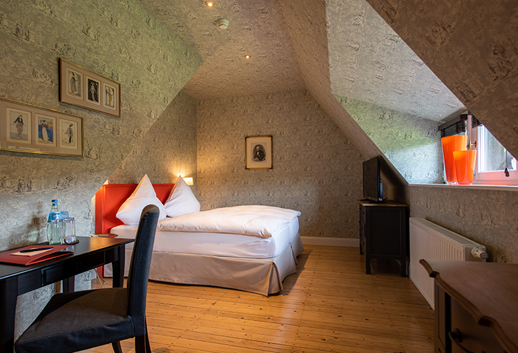 Dieses gemütliche Schlafzimmer im Dachgeschoss kombiniert Logis mit Liebe zum Detail mit Dachschräge, Doppelbett, Schreibtisch, Stuhl und Fernseher auf Holzboden. Der Raum zelebriert alte Baukultur durch seine gerahmten Bilder und ein Fenster mit Jalousien, die Charme und Eleganz ausstrahlen.