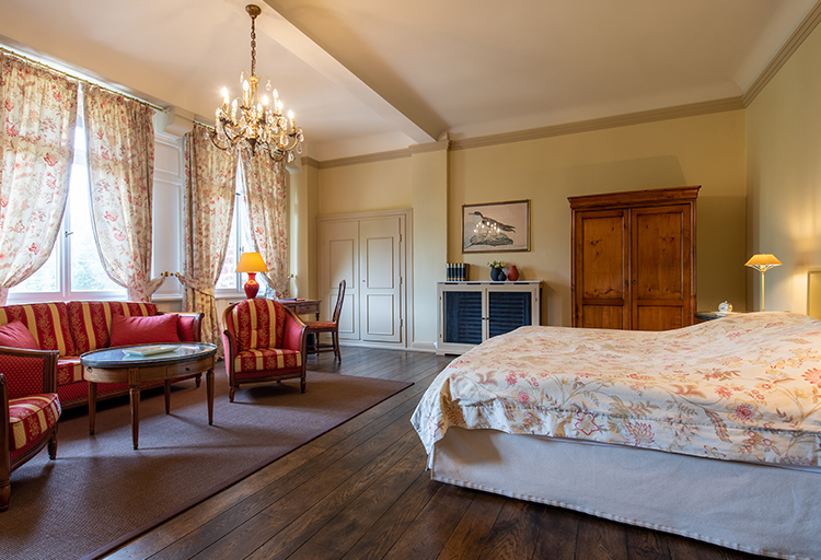 Ein geräumiges Zimmer mit einem geblümten Bett, einer roten Sofagarnitur, einem Kronleuchter und einem Holzboden strahlt Behaglichkeit aus. Der Kleiderschrank und die großen Fenster, die mit geblümten Vorhängen geschmückt sind, runden das gemütliche Ambiente ab.