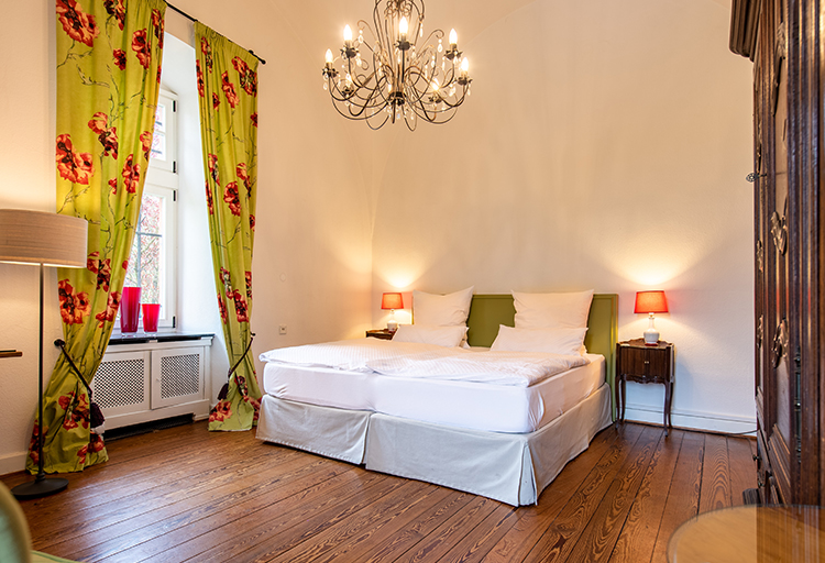 Dieses elegant eingerichtete Schlafzimmer, das an Schloss Saareck erinnert, verfügt über ein Doppelbett mit grünem Kopfteil, geblümte Vorhänge, Holzboden, Kronleuchter und von der Baukultur inspirierte Elemente wie zwei Nachttische mit roten Lampen und einen großen Holzkleiderschrank.