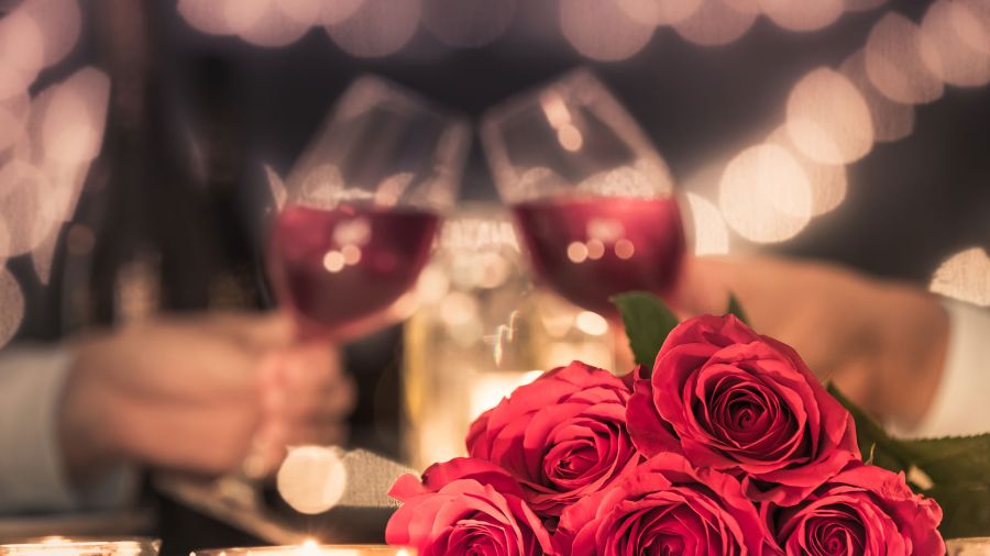 Close-up of red roses with two hands clinking glasses of red wine and blurred lights in the background.