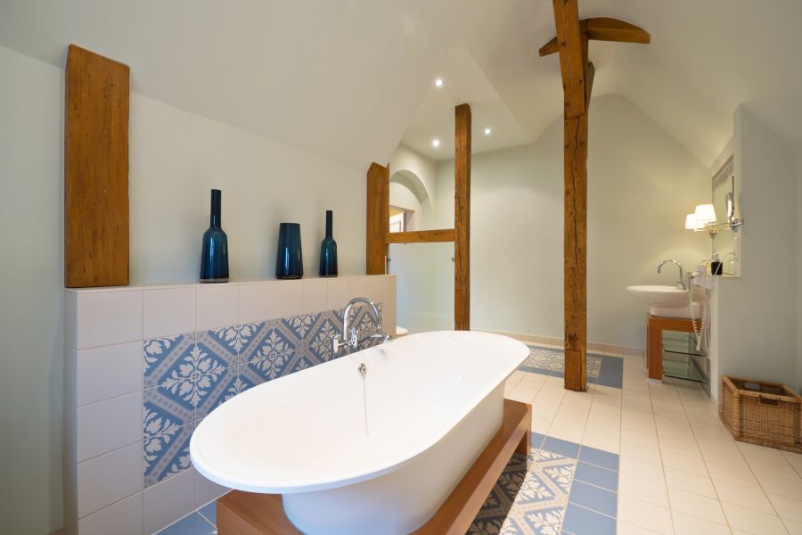 Großzügiges Badezimmer mit weißer freistehender Badewanne, dekorativen blauen Fliesen, die an alte Baukultur erinnern, Holzbalken und blauen Vasen. Weißes Waschbecken und Weidenkorb sorgen im Hintergrund für charmante Annehmlichkeiten.