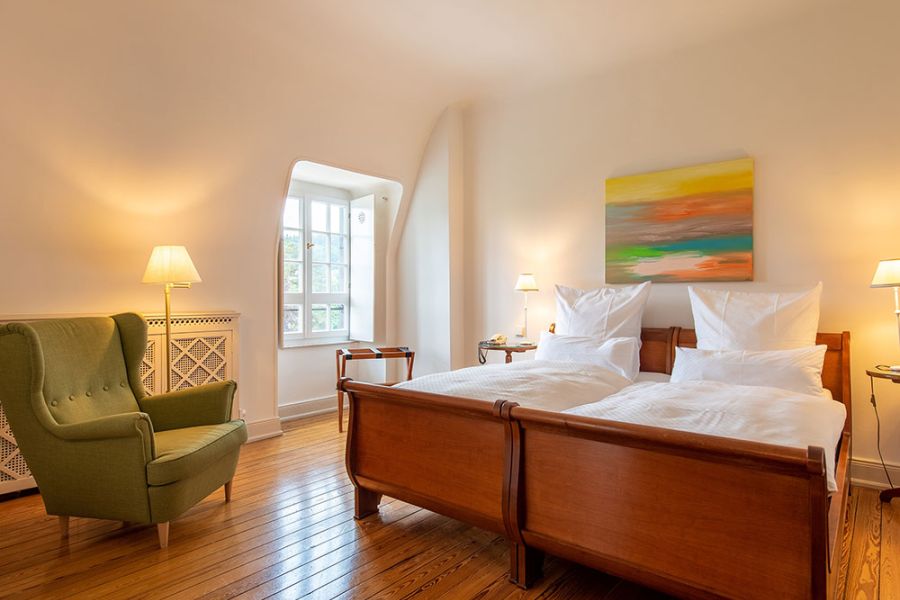 Ein gemütliches Schlafzimmer mit Doppelbett, grünem Sessel und warmer Beleuchtung verkörpert Behaglichkeit. Über dem Bett hängt ein farbenfrohes Gemälde, das an Baukultur erinnert, während Sonnenlicht durch ein Fenster mit weißem Rahmen strömt, das an den zeitlosen Charme von Schloss Saareck erinnert.