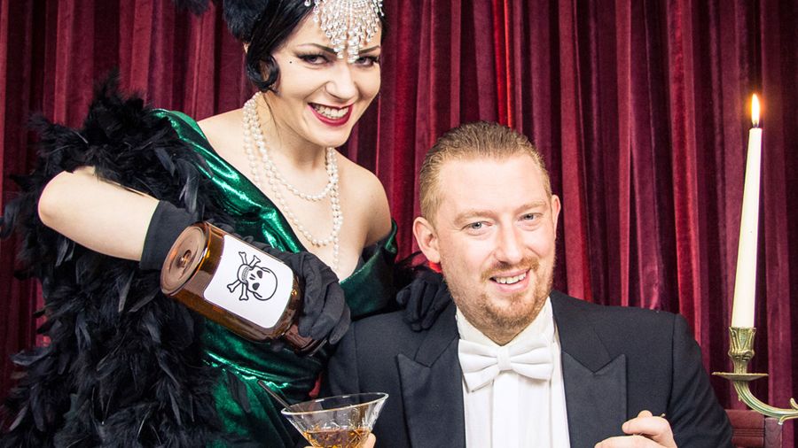 Glamouröse Darstellerin im 20er-Jahre-Kostüm mit grünem Kleid und Federboa gießt heimlich Gift aus einer Flasche mit Totenkopf in das Glas eines ahnungslosen Mannes im Smoking – vor rotem Samtvorhang bei Kerzenlicht