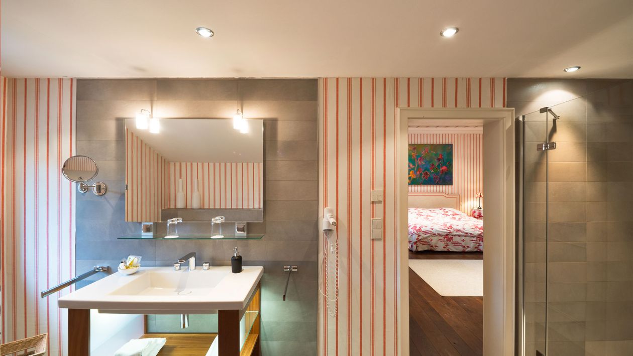 Modernes Badezimmer mit gestreifter Tapete, rechteckigem Spiegel, Waschbecken und ebenerdiger Dusche. Eine offene Tür gibt den Blick auf ein Schlafzimmer mit geblümter Tagesdecke frei, die an die Behaglichkeit der traditionellen alten Baukultur erinnert.