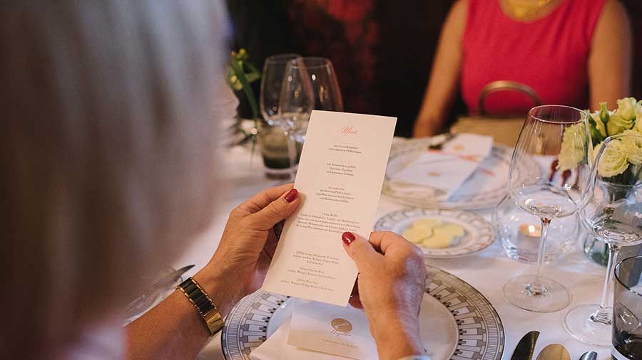 Eine Person entspannt sich an einem Restauranttisch und hält eine Speisekarte zwischen leeren Weingläsern und einem Brotteller mit Butter in der Hand, während ein Blumengesteck den Genuss des Augenblicks steigert.