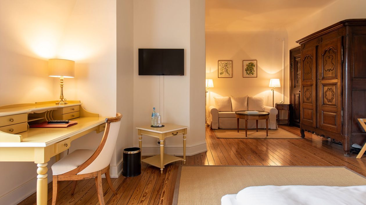 Das warm beleuchtete Hotelzimmer bietet hervorragende Annehmlichkeiten mit einem Schreibtisch, einem Stuhl und einem kleinen an der Wand montierten Fernseher. Ein Sitzbereich verfügt über ein Sofa und einen großen Holzschrank. Hartholzböden und sanfte Beleuchtung verstärken das gemütliche Ambiente und spiegeln die zeitlose Eleganz der Baukultur wider.