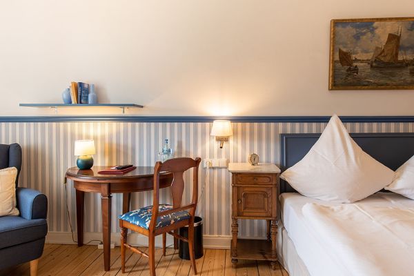 Gemütliches Hotelzimmer mit Bett, Nachttisch und Holzschreibtisch mit Stuhl. Zwei Lampen sorgen für warmes Licht. Ein Gemälde und Bücher auf einem Regal bereichern das Dekor und spiegeln den Charme der lokalen Baukultur wider. Genießen Sie moderne Annehmlichkeiten in dieser kulturellen Oase in der Nähe von Schloss Saareck.