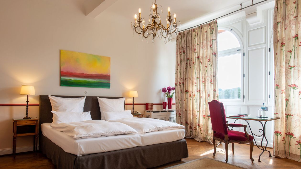 Das warm beleuchtete Hotelzimmer bietet hervorragende Annehmlichkeiten mit einem Schreibtisch, einem Stuhl und einem kleinen an der Wand montierten Fernseher. Ein Sitzbereich verfügt über ein Sofa und einen großen Holzschrank. Hartholzböden und sanfte Beleuchtung verstärken das gemütliche Ambiente und spiegeln die zeitlose Eleganz der Baukultur wider.