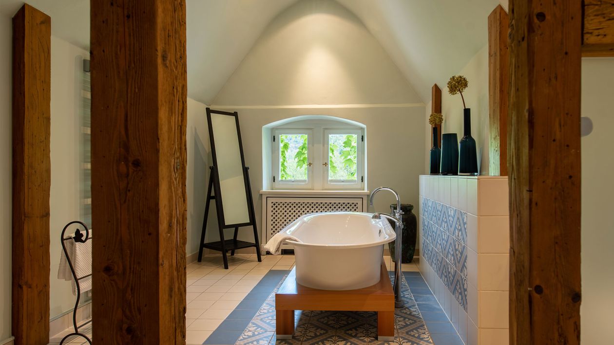 Ein Badezimmer mit baukulturellen Designelementen, einer freistehenden Badewanne, Holzbalken, einer Handtuchleiter, dekorativen Vasen und einem Fenster mit Blick auf üppiges Grün.