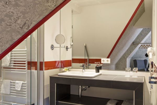 Dieses moderne Badezimmer im Stil der Baukultur von Schloss Saareck verfügt über einen großen Spiegel, ein Waschbecken und einen an der Wand montierten Haartrockner. Es verfügt über eine schräge Decke, eine Dusche und Handtücher auf einem beheizten Ständer. Dezente rote und graue Akzente verleihen diesem stilvollen Logis-Erlebnis Charme.