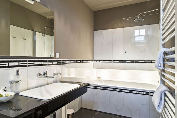 Ein modernes Badezimmer im Schloss Saareck, in dem alte Baukultur auf zeitgenössisches Design trifft. Es verfügt über ein wandmontiertes Waschbecken, eine Badewanne und eine Kopfbrause mit weißen und schwarzen Fliesen. Ein Handtuchhalter mit Handtüchern und ein Spiegel über dem Waschbecken vervollständigen dieses Logis mit Liebe zum Detail.