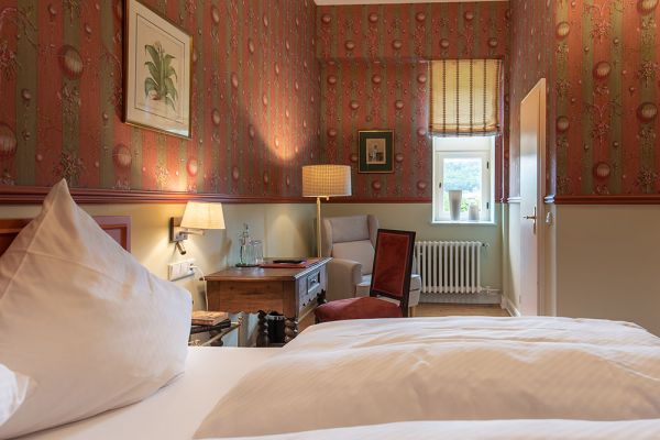 Ein gemütliches Zimmer mit gemusterter Tapete, einem Bett und einem hölzernen Nachttisch mit Lampe bietet Annehmlichkeiten, die an Schloss Saareck erinnern. Ein Sessel steht neben dem mit einer gemusterten Jalousie geschmückten Fenster und verbindet Komfort mit klassischer Baukultur.