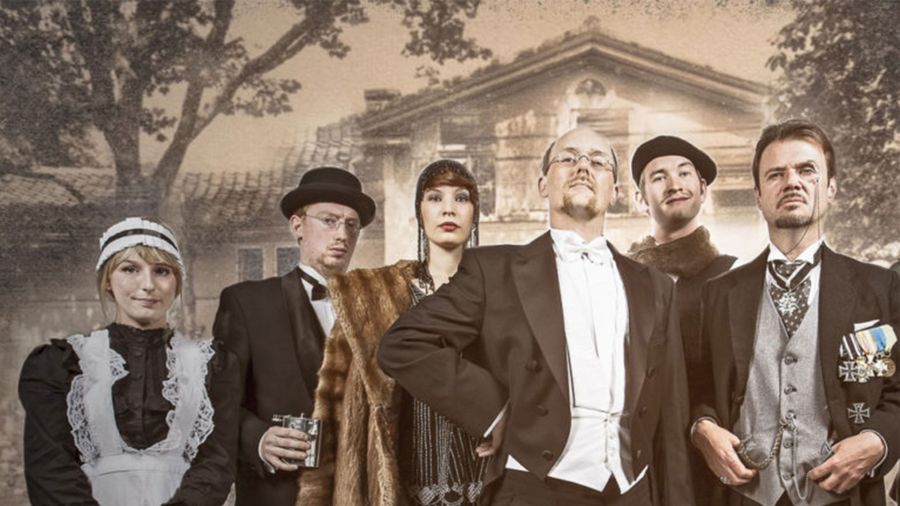 Six people in historical clothing in front of a large house, including a woman in a maid uniform and men in suits with bow ties, hats, and medals.