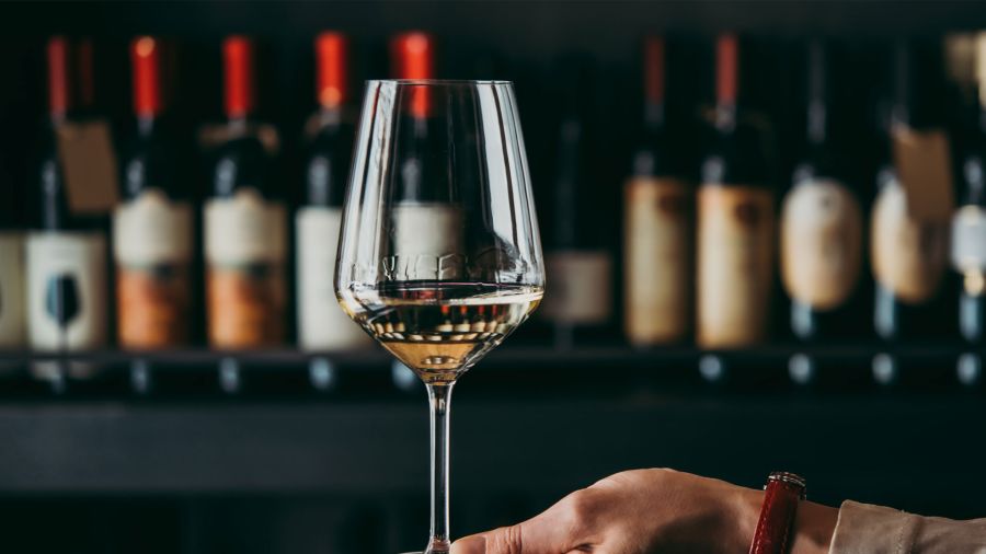 Ein gefülltes Weinglas mit Weißwein im Vordergrund, im Hintergrund eine unscharfe Reihe von Weinflaschen mit roten Kapseln auf einem Weinregal.