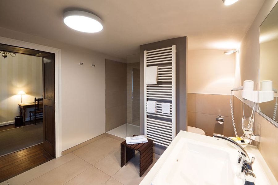 Dieses moderne Badezimmer im Stil der Baukultur verfügt über eine ebenerdige Dusche, einen weißen Handtuchhalter und einen großen Spiegel. Neben der Dusche steht eine Holzbank, die den Blick in den angrenzenden Raum mit Schreibtisch und Lampe freigibt – ein ruhiges Logis, das von zeitlosen Designprinzipien inspiriert ist.