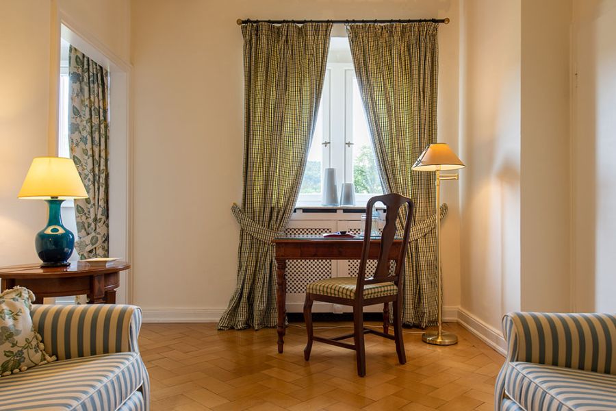 Ein Holzschreibtisch und ein Stuhl stehen an einem Fenster mit karierten Vorhängen, was an die zeitlose Baukultur von Schloss Saareck erinnert. Zwei Tischlampen und Sessel mit gestreiftem Stoff tragen zur Gemütlichkeit bei, während eine Stehlampe den Holzboden daneben schmückt.