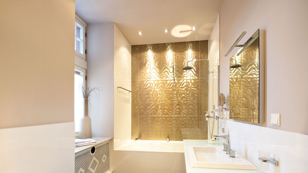 Modernes Badezimmer in beiger Farbgebung und im Sinne der Baukultur mit weißer Badewanne, strukturierter Wandverkleidung, weißem Waschbecken, Spiegel und Fenster, das Tageslicht hereinlässt.