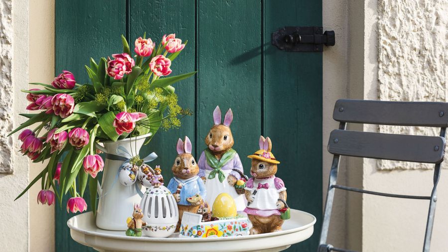 A white table with bunny figurines, a vase of pink tulips and various Easter decorations creates a delightful scene reminiscent of the elegant charm of Saareck Castle. A green door and a grey chair complete the enchanting setting.